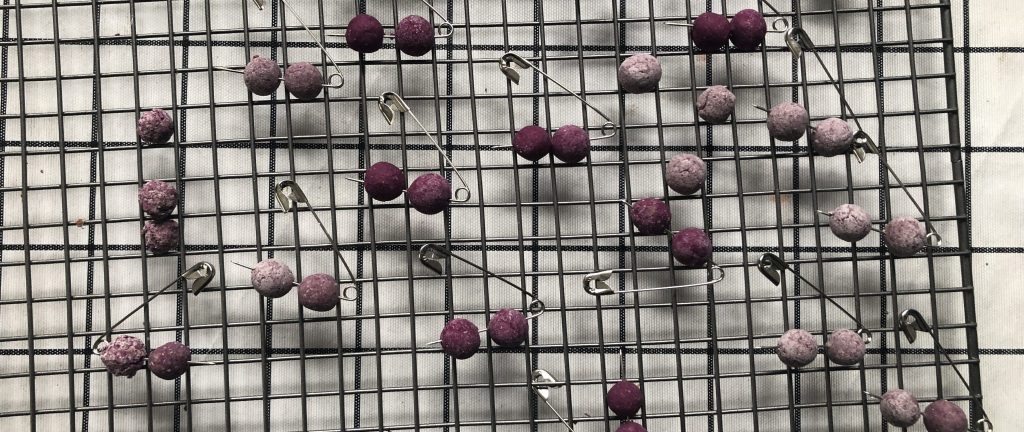 Light grey and purple beads, impaled on safety pins on a drying rack.