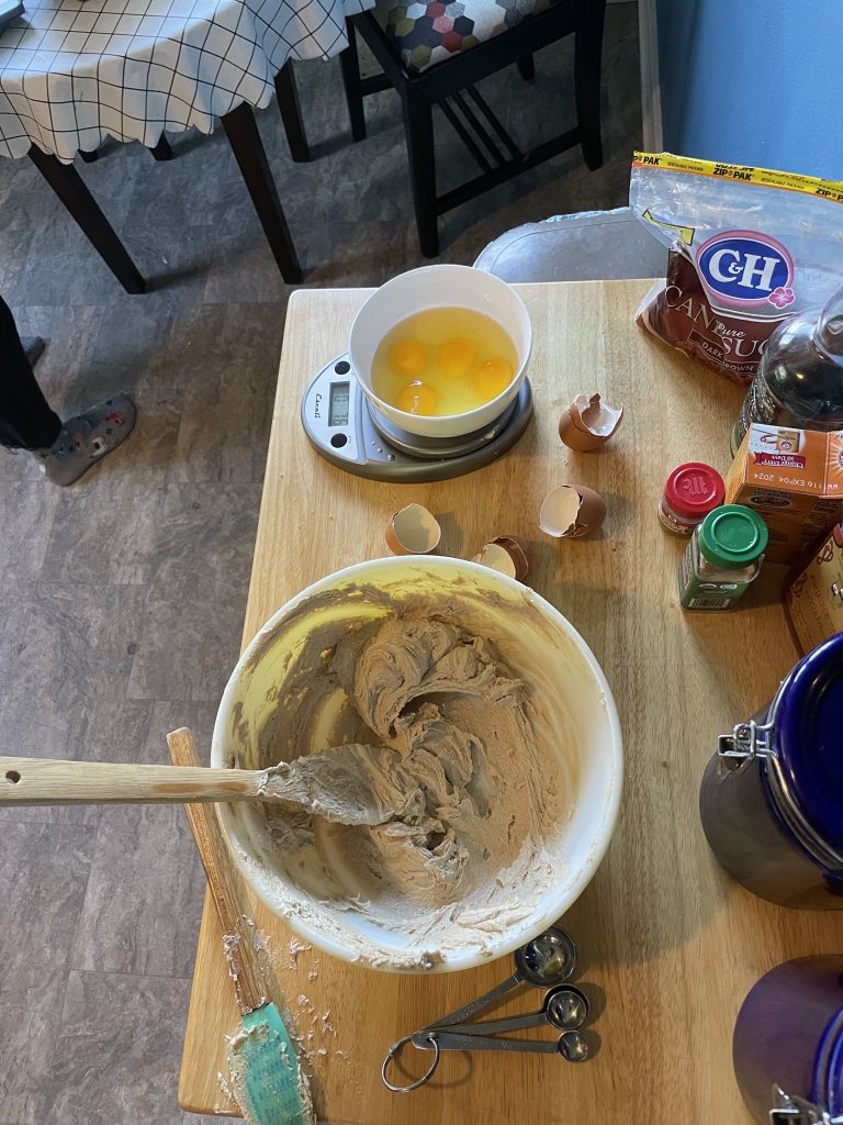 a messy countertop (egg shells, spoons, spice containers) with a yellow bowl with creamed butter and sugar in it, a bowl of eggs on a scale sits next to it.