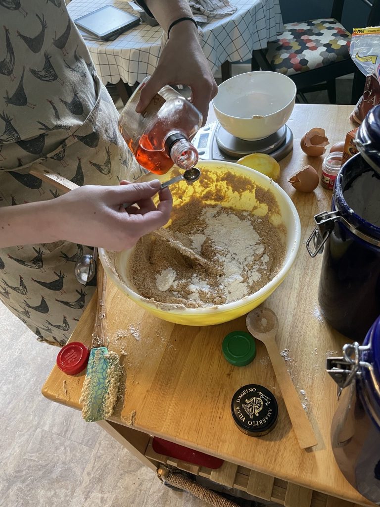A messy countertop (egg shells, spoons, spice containers) with a yellow bowl with cake mix in it, a woman is measuring amaretto to add to the bowl.