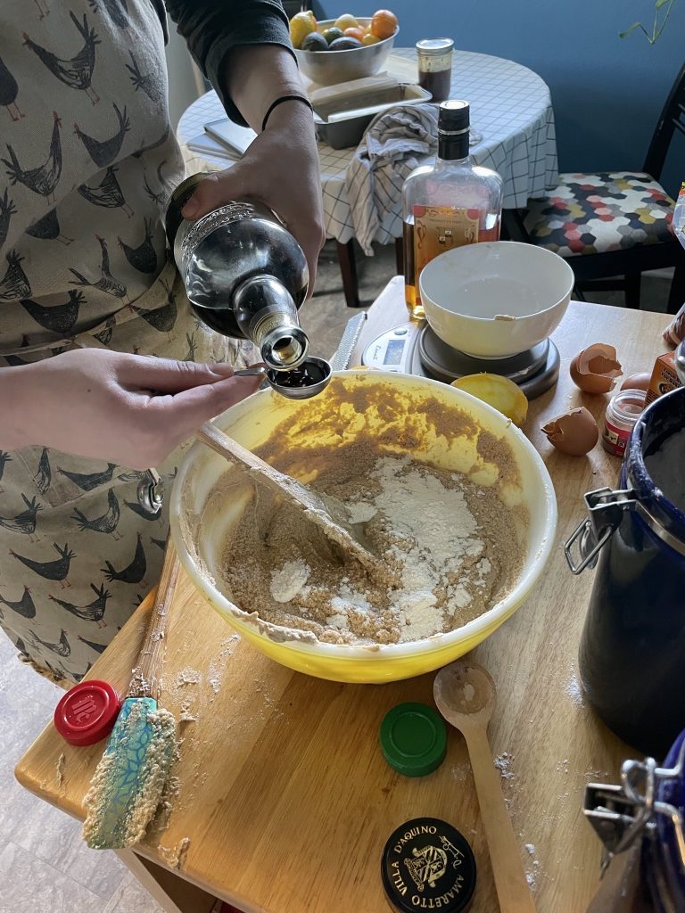 a messy countertop (egg shells, spoons, spice containers) with a yellow bowl with cake mix in it, a woman is measuring dark rum to add to the bowl.