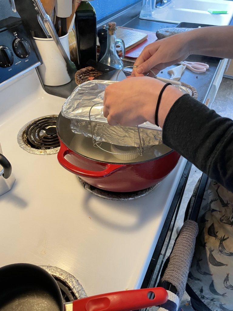 A woman uses the handles of a steam tray to lower a loaf pan wrapped in tinfoil into a red dutch oven.