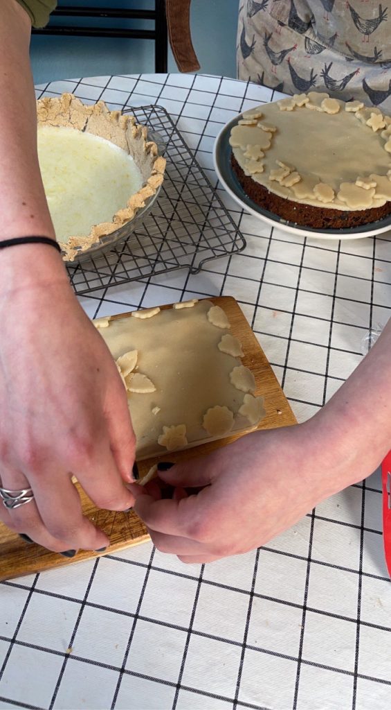 A round table with pie and two fruit cakes, one round, one loaf. A woman's hands are affixing an almond paste decoration to the side of the loaf.