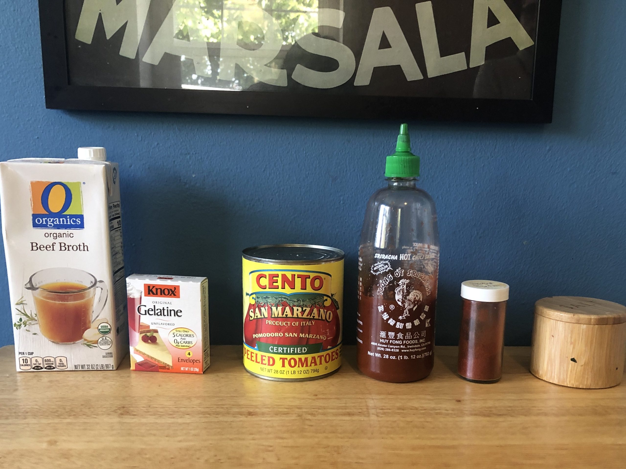 A wooden counter with beef broth, gelatin, canned tomatoes, sriracha, paprika, and salt.