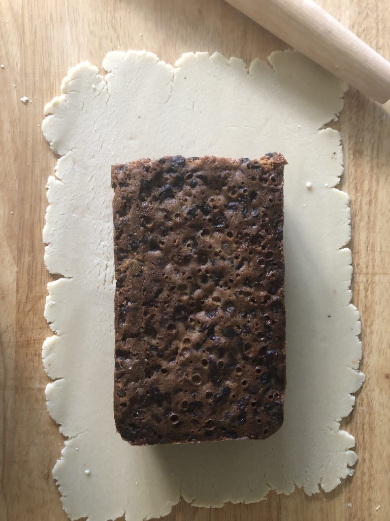 A rolled out rectangle of almond paste with a loaf of fruit cake on top of it.