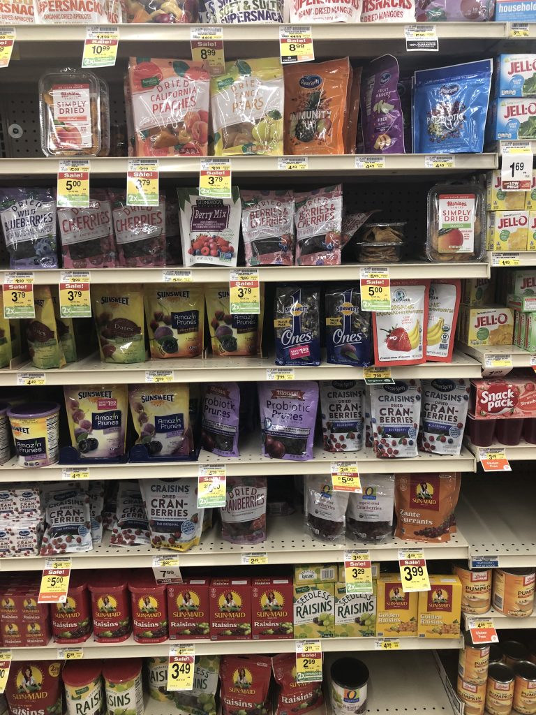 Shelves of dried fruit at a grocery store, packages are plastic, colorful, with lots of text, prices and sale signs are visible.
