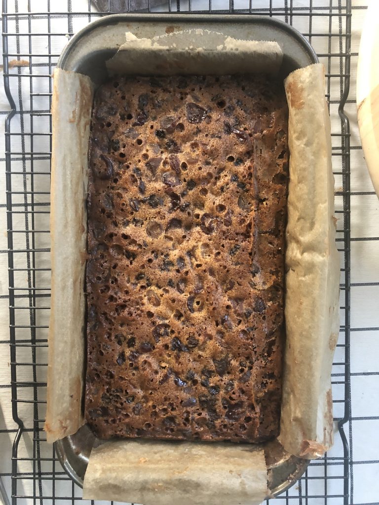 A dark brown fruit cake in a loaf pan. The top of the cake has lots of steam holes in it.